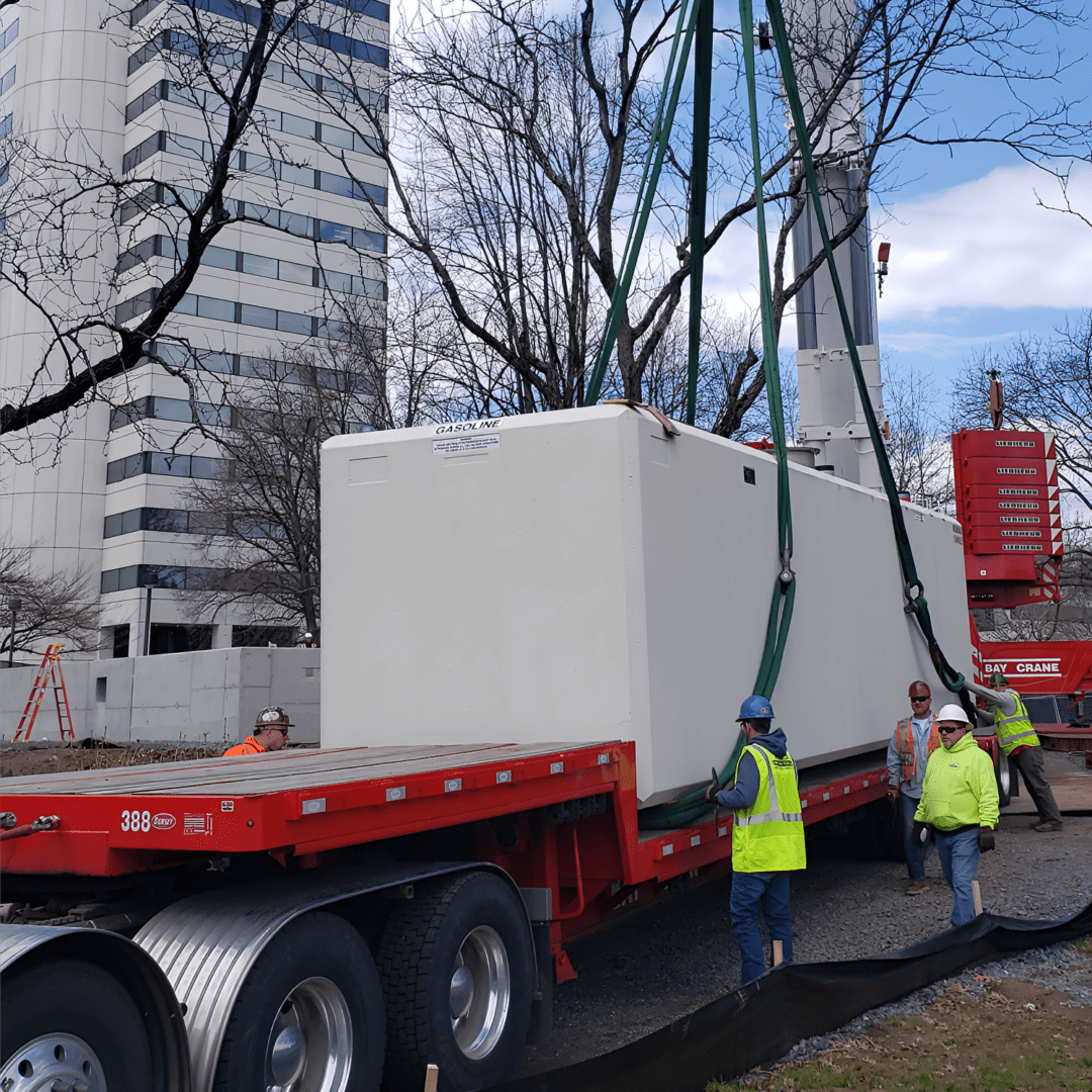 Workers guide a large white container being lifted by a crane onto a flatbed truck at a construction site near tall buildings.